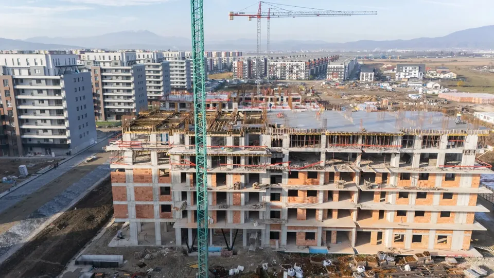 Un șantier cu un schelet de beton și cărămizi de culoare deschisă este vizibil sub un manipulator de culoare verde și un macarale roșu.