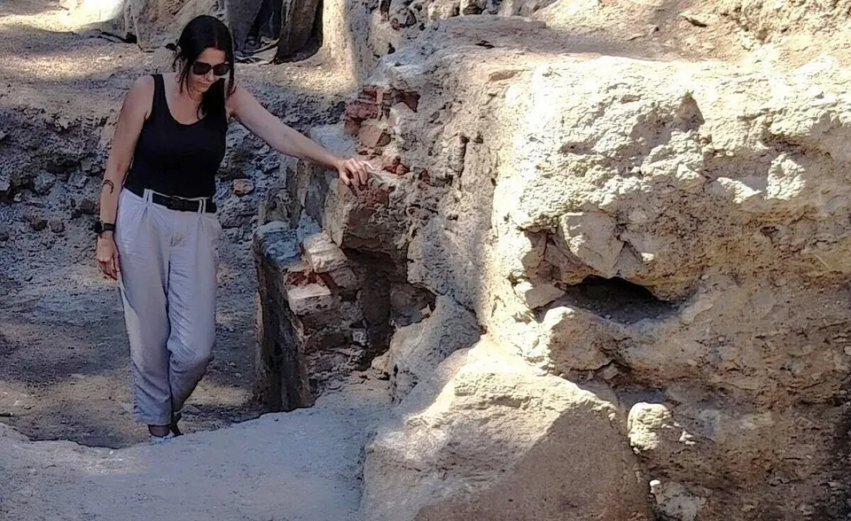 O femeie cu pantaloni deschiși și canotieră neagră stă lângă un zid de piatră și cărămidă într-o zonă de săpătură arheologică.