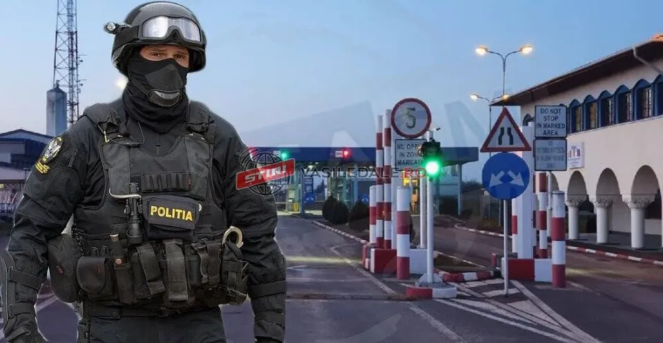 Un agent de poliție în echipament tactice negru, cu cască și mască, stă în prim-plan în fața unui punct de trecere a frontierei cu semne rutiere.