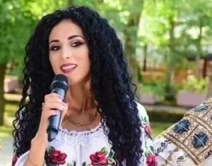 O femeie cu părul negru și crepus, purtând o bluză albă cu motive florale, ține un microfon negru în mână.