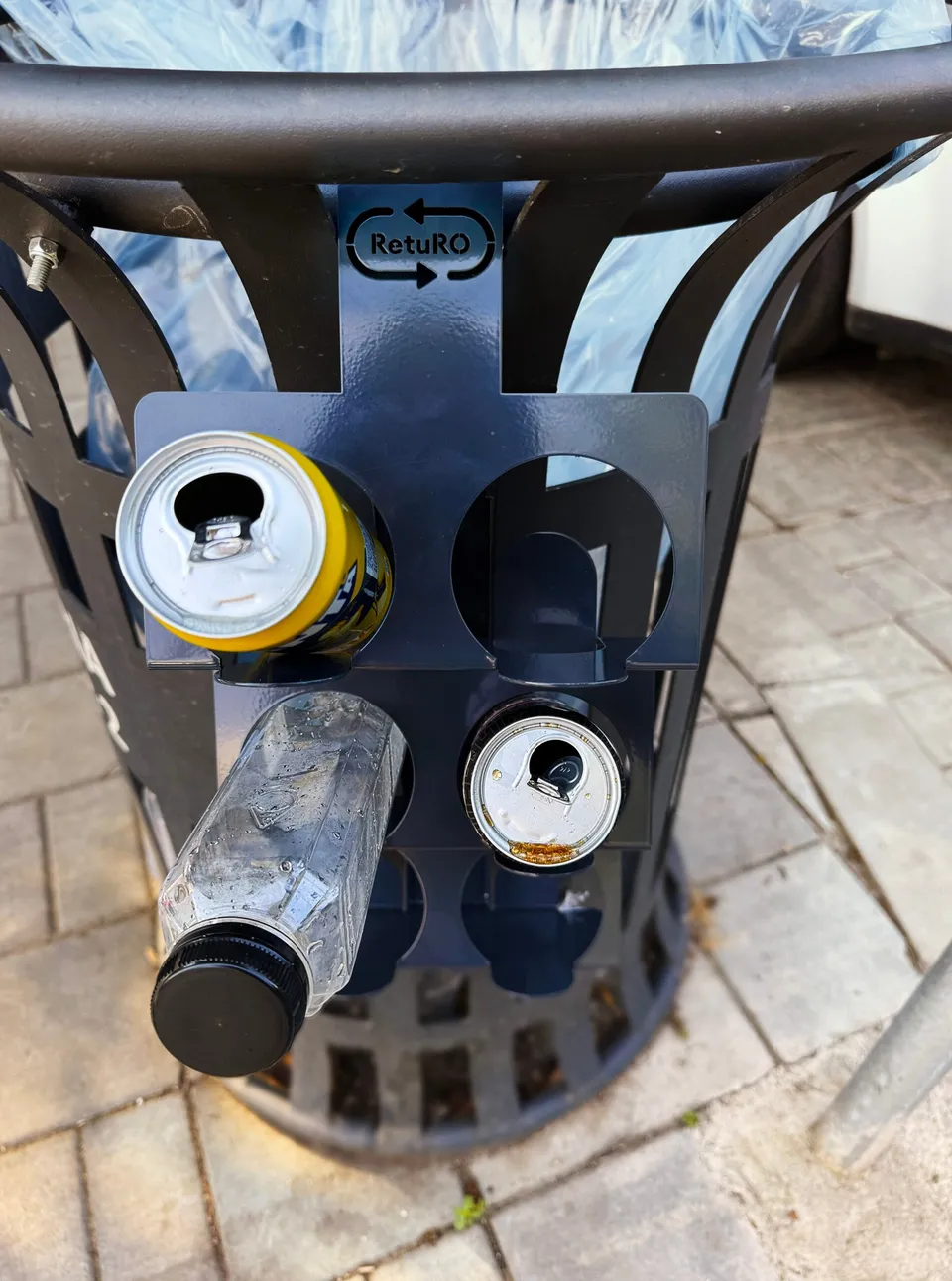 Un coș de gunoi metalic negru cu orificii circulare în care sunt introduse o conservă de aluminiu și o sticlă de plastic.