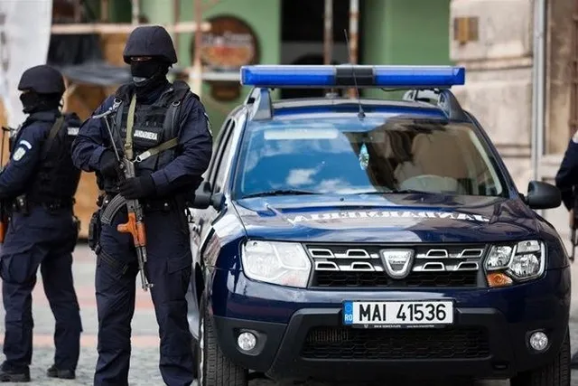 Două persoane în uniforme de jandarm și măști negre stau lângă un autovehicul albastru cu inscripții și bară de lumini albastră.