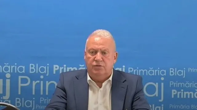 Un bărbat cu părul scurt și alb, purtând un sacou albastru și cămașă albă, stă în fața unui fundal albastru cu text repetat.