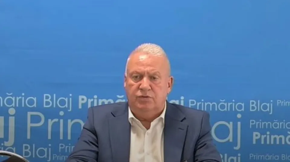 Un bărbat cu părul scurt și alb, purtând un sacou albastru și cămașă albă, stă în fața unui fundal albastru cu text repetat.