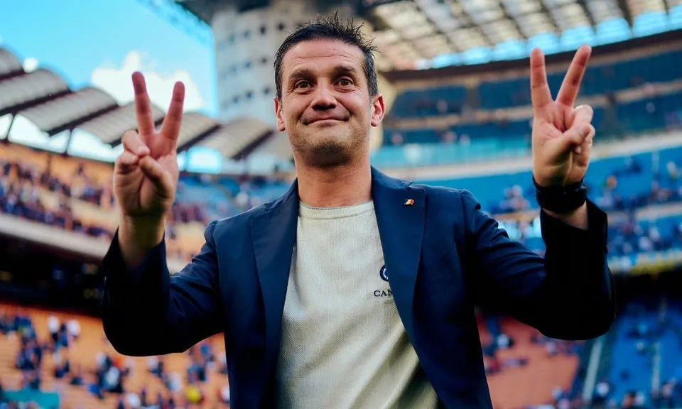 Un bărbat cu sacou închis și tricou deschis la culoare face semnul victoria cu ambele mâini în fața unui stadion plin de oameni.