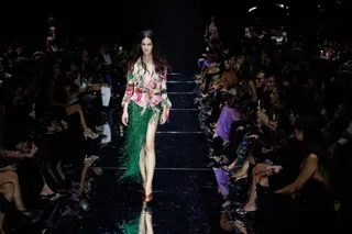 O femeie cu părul lung merge pe o pistă de catwalk purtând o bluză cu imprimeu floral și o fustă lungă, verde, cu margini de infilări.