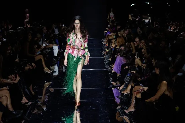 O femeie cu părul lung merge pe o pistă de catwalk purtând o bluză cu imprimeu floral și o fustă lungă, verde, cu margini de infilări.