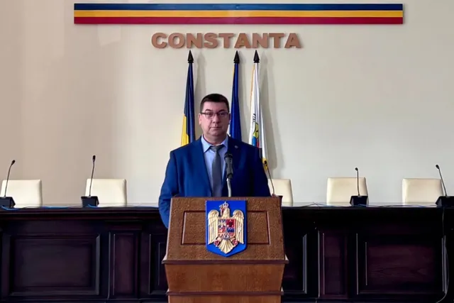 Un bărbat cu ochelari și costum albastru stă în spatele unui podium de lemn pe care este afișat un scut colorat.