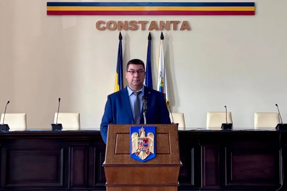 Un bărbat cu ochelari și costum albastru stă în spatele unui podium de lemn pe care este afișat un scut colorat.