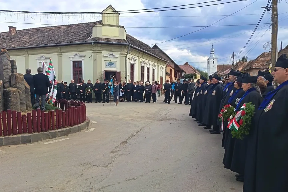 Un grup de persoane îmbrăcate în haine negre stă în rând pe o stradă lângă un monument și o clădire cu acoperiș înclinat.