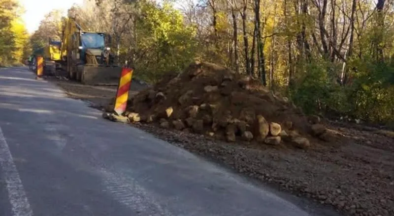 Un utilaj de construcții și un morman de pământ cu bucăți de lemn sunt situate pe marginea unui drum asfaltat lângă o pădure.