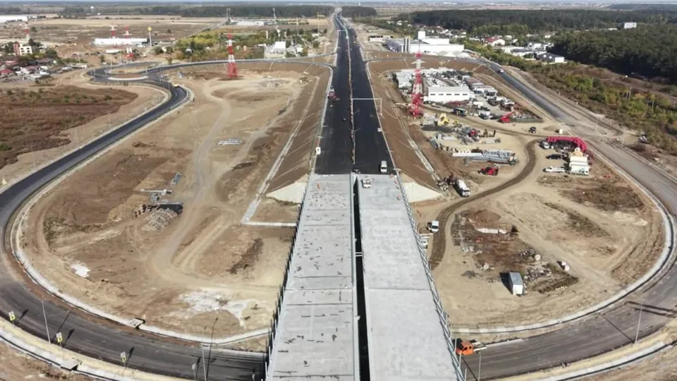 O vedere aeriană prezintă o autostradă cu asfalt negru, un viaduct de beton și drumuri de acces în curs de construcție pe un teren de pământ.