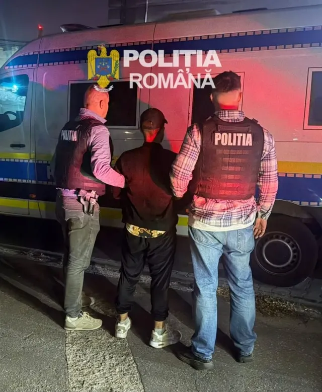 Trei persoane, dintre care doi polițiști în uniformă și un bărbat cu mâinile în spate, stau lângă un vehicul alb cu inscripții de poliție.