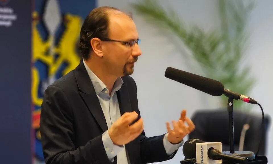 Un bărbat cu ochelari și sacou închis, purtând o cămașă deschisă la guler, vorbește în fața unui microfon.