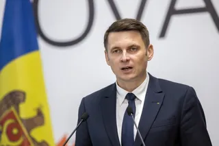 Un bărbat cu părul scurt, îmbrăcat în costum închis și cravată, stă în fața unui fundal alb și a unui steag colorat lângă microfoane.