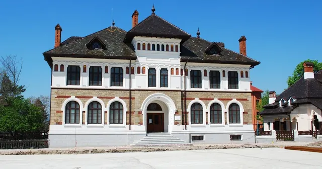 O clădire cu fațadă albă și brună, acoperiș în șarpă și ferestre arcuite, este situată sub un cer senin.