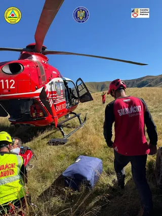Un elicopter roșu cu inscripția SMURD stă pe o pajiță de munte lângă persoane purtând veste reflectorizante și echipament de protecție.