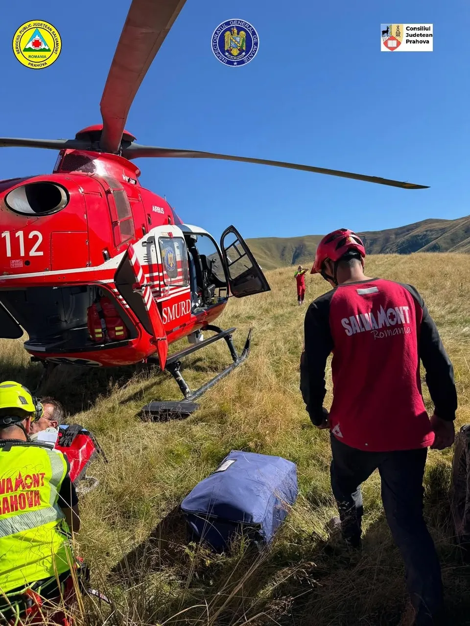 Un elicopter roșu cu inscripția SMURD stă pe o pajiță de munte lângă persoane purtând veste reflectorizante și echipament de protecție.