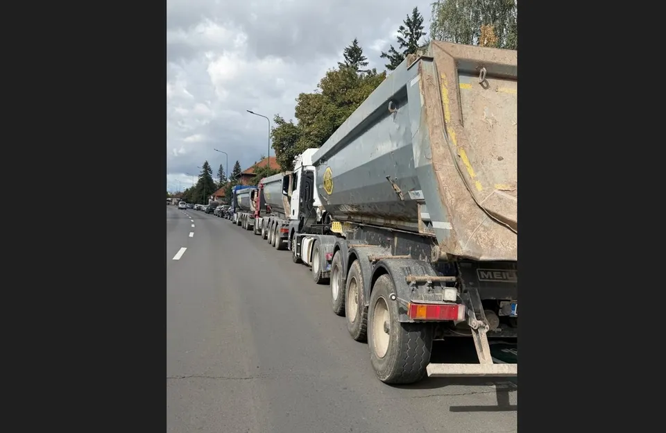 O serie de autoutilitare și camioane cu remorci sunt parcate pe marginea unui drum asfaltat sub un cer înnorat.
