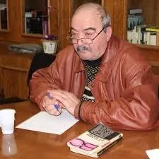 Un bărbat cu ochelari și o jachetă de piele maro scrie pe o foaie de hârtie de la o masă de lemn.