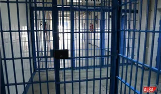 Gratiile albastre ale unei celule de detenție se deschid spre un coridor cu pereți albi și plăci de faianță.