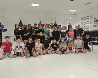 Un grup de persoane posedă diverse ținute sportive și stau grupate pe un covor alb într-o sală de antrenament cu saci de box suspendați.