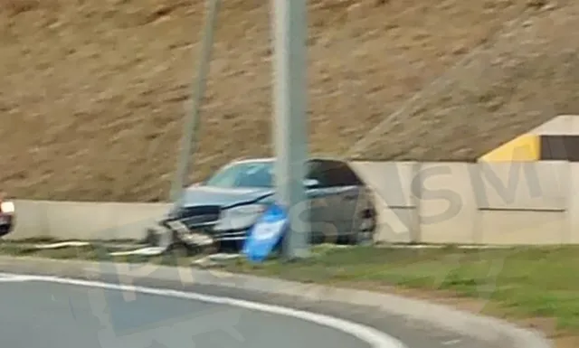 O mașină gri este parcată pe marginea unui drum lângă un gard de beton și un taluz de pământ.