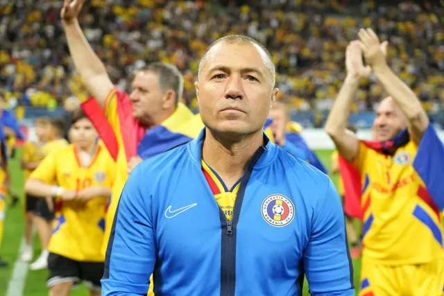 Un bărbat cu părul scurt și haine sport albastre stă în prim-plan, în timp ce alți oameni în echipamente galbene aplaudă în fundalul unui stadion.