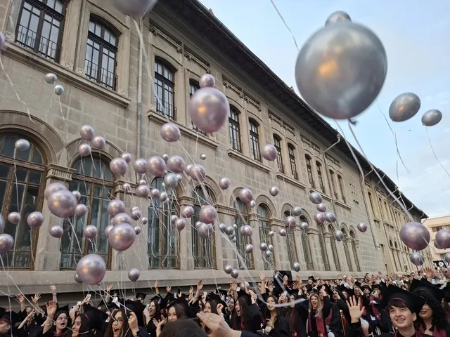 O mulțime de persoane poartă haine negre și lanțuri colorate, lansând numeroase baloane argintii în fața unei clădiri înalte cu ferestre mari.