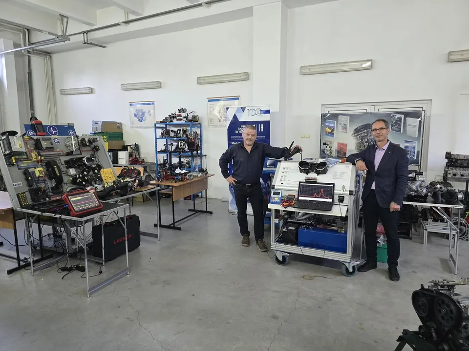 Doi bărbați stau într-un laborator lângă un echipament tehnic pe cărucior și mese cu diverse dispozitive electronice și componente mecanice.