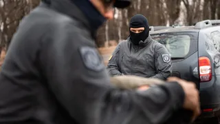 Un bărbat cu mască neagră și uniformă gri stă lângă un autoturism gri, în timp ce un alt individ în uniformă este în prim-plan.