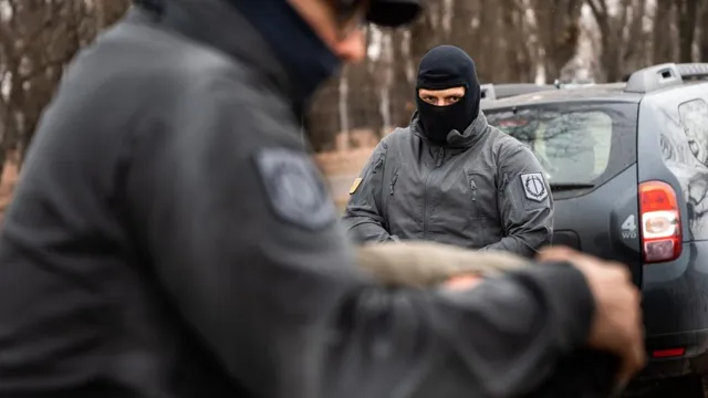 Un bărbat cu mască neagră și uniformă gri stă lângă un autoturism gri, în timp ce un alt individ în uniformă este în prim-plan.