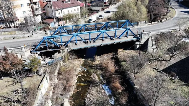 O structură metalică albastră de tip pod traversează un râu îngust, fiind situată între o zonă rezidențială și un drum asfaltat.