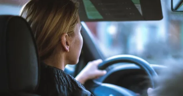 O femeie cu părul blond este așezată pe scaunul șoferului, ținând volanul unui vehicul.