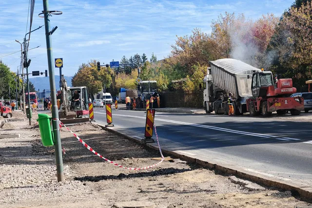 Un camion cu benă ridicată și utilaje de construcții sunt poziționate pe o stradă asfaltată lângă un trotuar în curs de renovare.