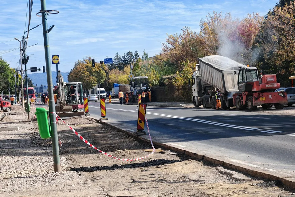 Un camion cu benă ridicată și utilaje de construcții sunt poziționate pe o stradă asfaltată lângă un trotuar în curs de renovare.