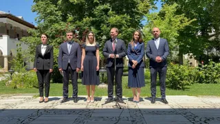 Șase persoane îmbrăcate în haine formale stau în rând pe un teren împădurit în fața unei clădiri albe.