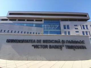 O clădire modernă cu fațadă gri și panouri de sticlă este situată în spatele unui zid pe care scrie un text în litere închise la culoare.