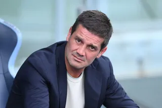 Un bărbat cu părul scurt și întunecat poartă un sacou albastru peste un tricou alb și privește spre cameră.