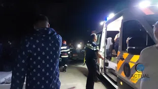 Un bărbat în cămașă albastră cu modele albe stă cu spatele, lângă o ambulanță cu portierele deschise și un oficial în uniformă întunecată.