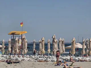 Pe o plajă însorită, șezlonguri și umbrele albe sunt așezate în rânduri, iar câțiva oameni stau întinși sau în picioare lângă mare.