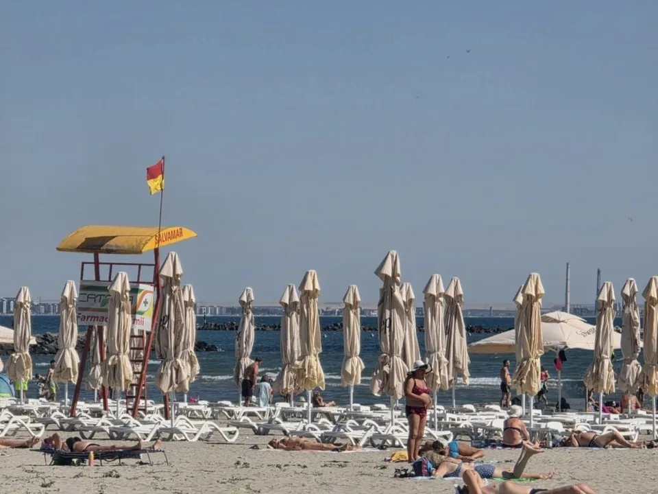 Pe o plajă însorită, șezlonguri și umbrele albe sunt așezate în rânduri, iar câțiva oameni stau întinși sau în picioare lângă mare.