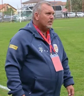 Un bărbat cu barbă scurtă poartă un hanorac albastru cu logo-uri și un card de identificare suspendat la gât pe un teren de fotbal.