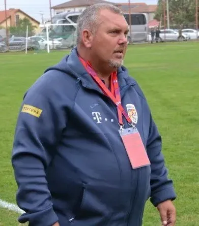 Un bărbat cu barbă scurtă poartă un hanorac albastru cu logo-uri și un card de identificare suspendat la gât pe un teren de fotbal.