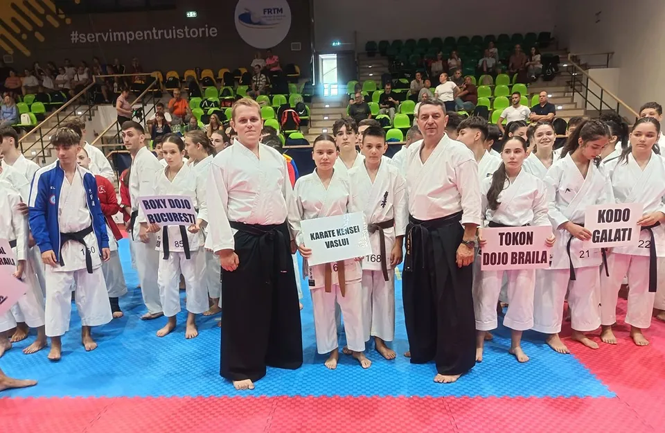 Un grup de sportivi în kimono-uri de karate, unii ținând pancarte cu numele cluburilor, stau pe un tatami albastru și roșu, într-o sală cu tribune pline.