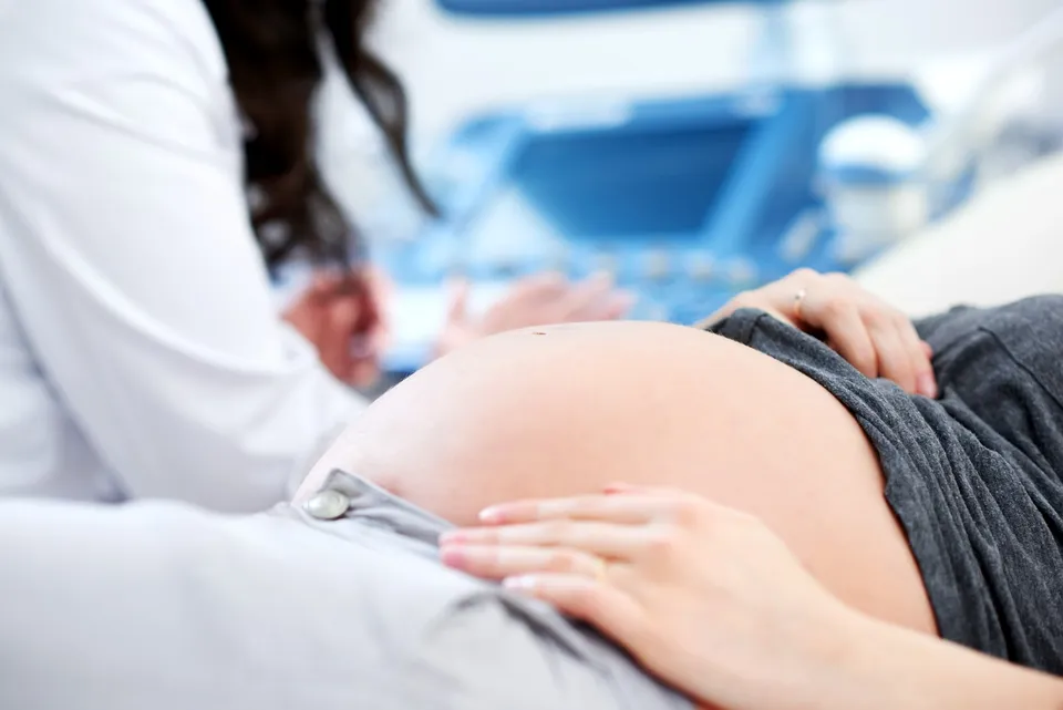 O persoană cu mâinile pe un abdomen proeminent poartă pantaloni gri, fiind așezată lângă o altă persoană îmbrăcată în alb.