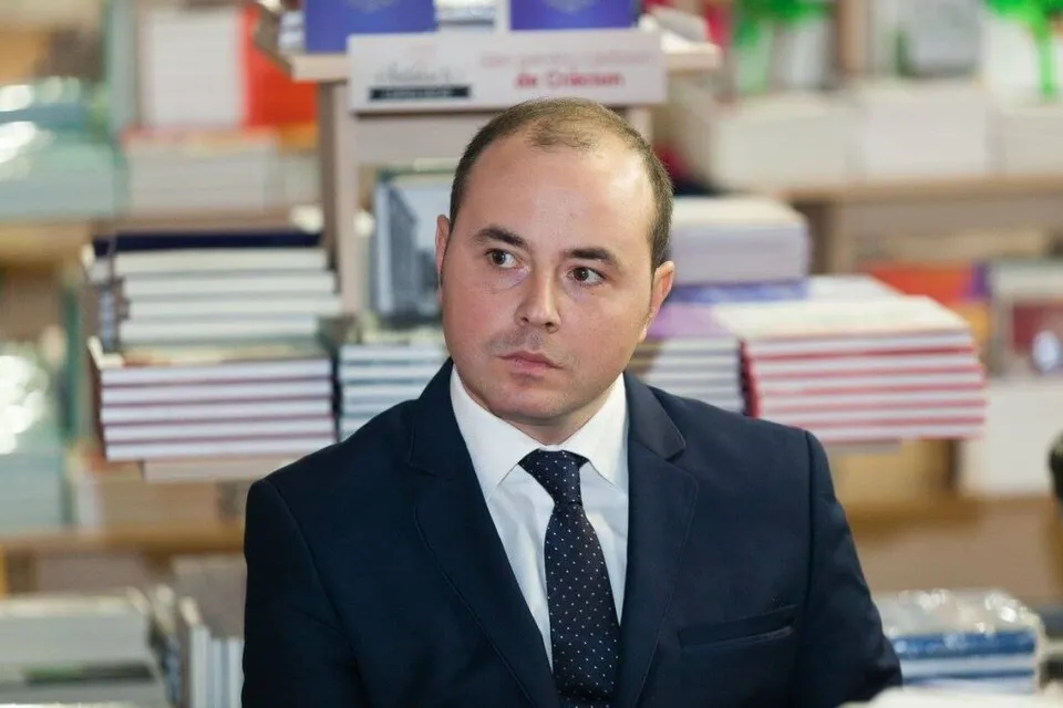 Un bărbat cu părul scurt și rare, purtând sacou închis la culoare, cămașă albă și cravată neagră cu puncte albe, privește spre lateral într-un decor cu rafturi.