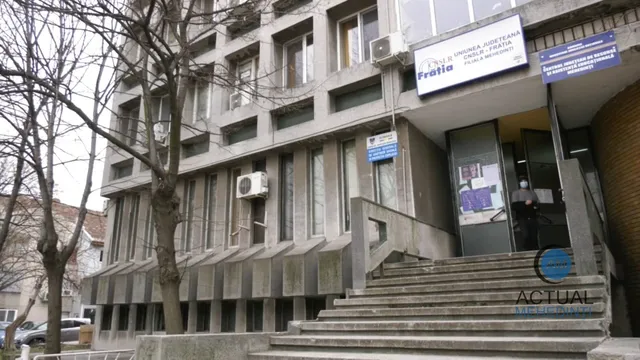 O clădire gri cu ferestre rectangulare, o scară de beton în prim-plan și un arbore fără frunze în partea stângă.