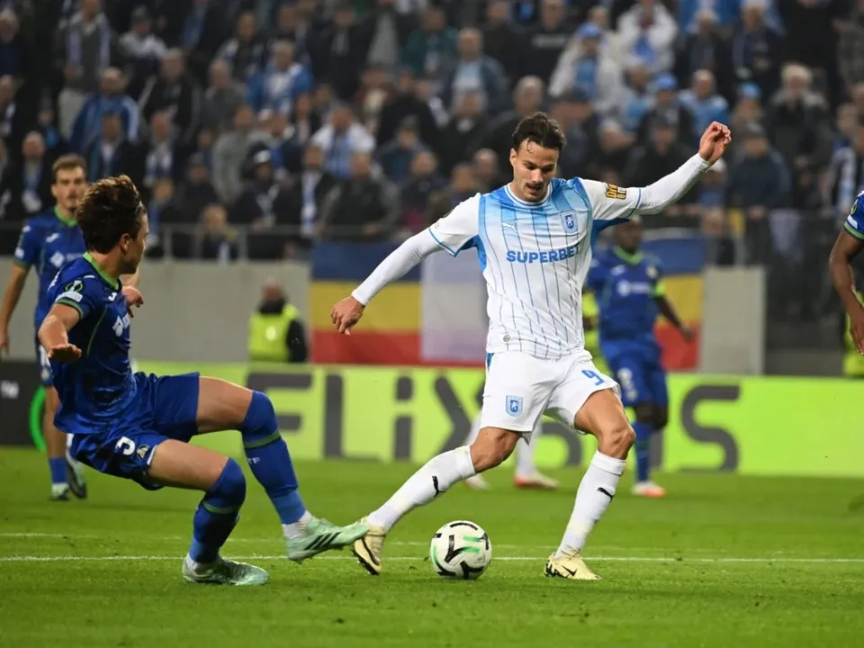 Un jucător în echipament alb și albastru conduce mingea pe terenul de fotbal în fața unui adversar în echipament albastru.
