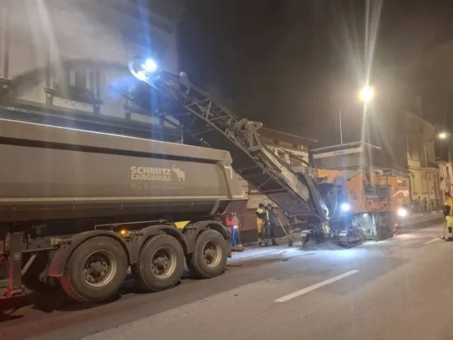 Un utilaj de construcții cu o bandă transportoare ridicate descarcă materiale într-un camion mare pe un drum asfaltat noaptea.